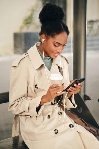 Atractiva chica afroamericana casual con elegante abrigo de trinchera y auriculares con café para ir felizmente usando el celular en la parada de autobús