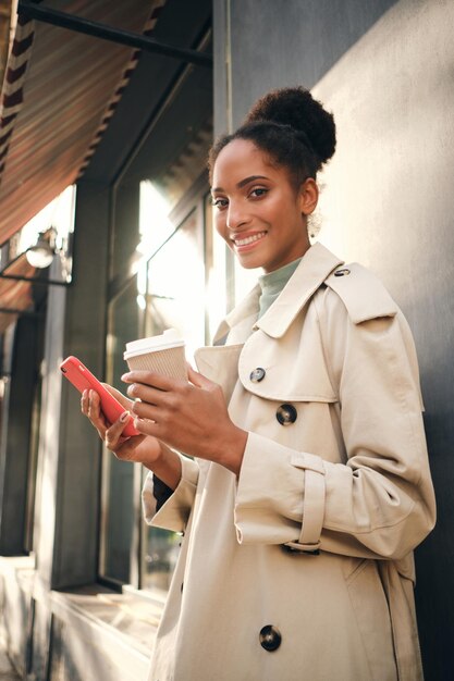 Atractiva chica afroamericana casual con elegante abrigo de trinchera con café para llevar y teléfono celular mirando alegremente a la cámara al aire libre