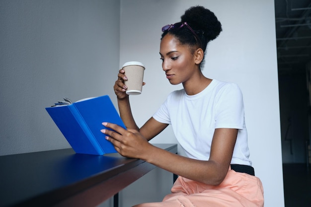 Atractiva chica afroamericana informal leyendo un libro y bebiendo café en un moderno espacio de trabajo conjunto