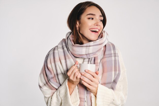 Atractiva chica alegre con una acogedora bufanda sosteniendo una taza con una bebida caliente felizmente mirando hacia otro lado sobre el fondo blanco