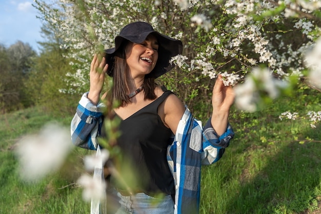 Atractiva chica alegre con un sombrero entre los árboles en flor en la primavera, en un estilo casual