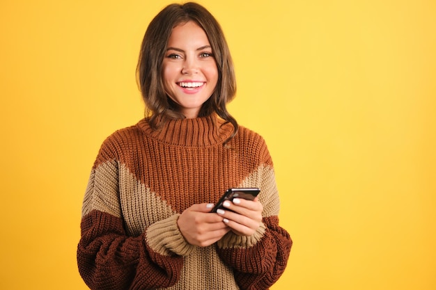 Atractiva chica alegre con suéter acogedor con celular mirando felizmente a la cámara sobre fondo amarillo