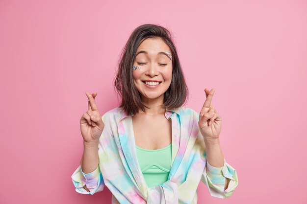 Foto gratuita atractiva chica asiática esperanzadora espera buena suerte mantiene los dedos cruzados cierra los ojos anticipa noticias positivas sonríe con alegría viste camisa colorida aislada sobre pared rosa. concepto de oración