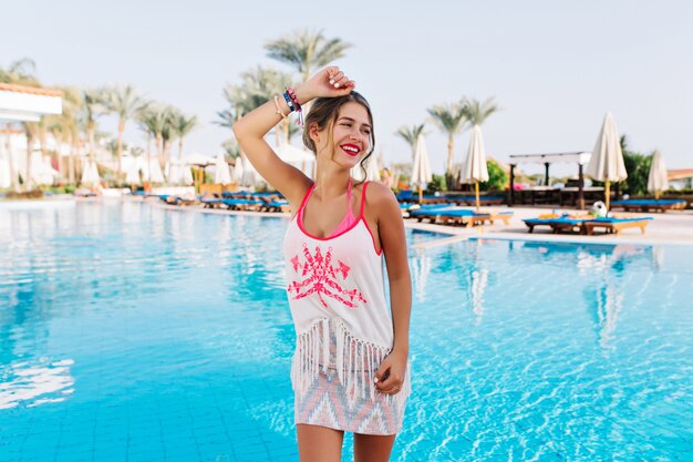 Atractiva chica bien formada con minifalda y camiseta sin mangas con flecos bailando con la mano arriba esperando divertirse en la piscina