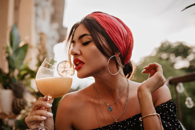 Atractiva chica de cabello moreno con diadema rosa aretes redondos de plata pañuelo brillante en camisa negra bebiendo cóctel