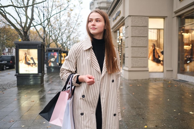 Atractiva chica casual caminando con confianza por la calle lluviosa de la ciudad con bolsas de compras