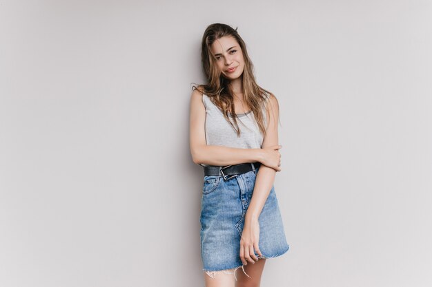 Atractiva chica caucásica en falda de mezclilla posando con placer. Foto interior de refinada mujer de cabello castaño con expresión de cara feliz aislada.