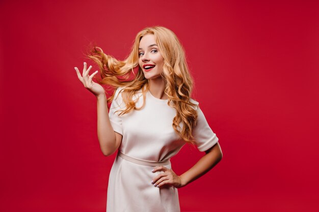 Atractiva chica caucásica jugando con su pelo largo en la pared roja. Foto interior de mujer elegante y atractiva en vestido.