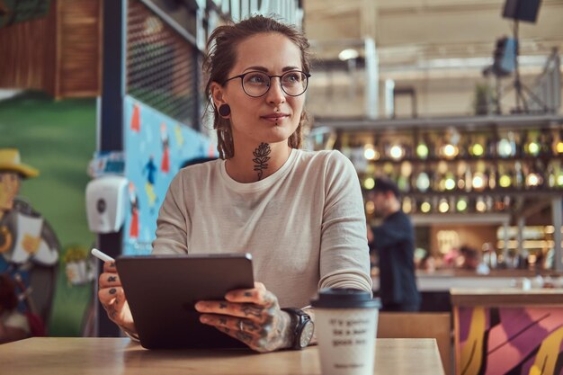 Atractiva chica creativa con tatuajes en las manos está sentada en el café mientras dibuja en su bloc de notas digital.