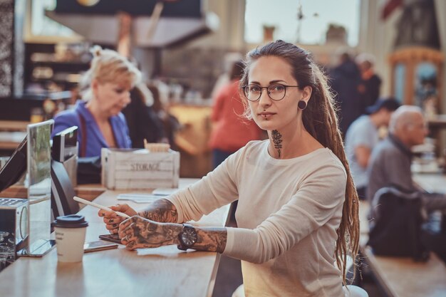 Atractiva chica creativa con tatuajes en las manos está sentada en el café mientras dibuja en su bloc de notas digital.