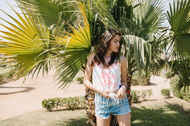 Atractiva chica delgada en pantalones cortos de mezclilla se esconde del sol bajo la palmera con expresión seria. Adorable morena joven en camiseta sin mangas de moda descansando en el exótico parque de vacaciones