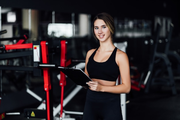 Atractiva chica deportiva, entrenadora personal en medio de un moderno gimnasio con un plan de entrenamiento en sus manos