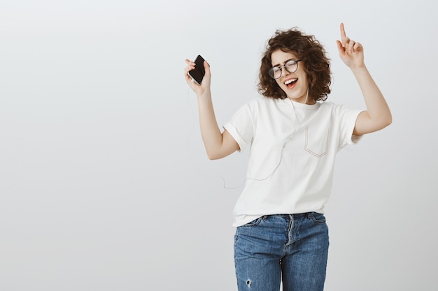 Atractiva chica despreocupada bailando con música en el teléfono móvil, escuchando canciones en auriculares