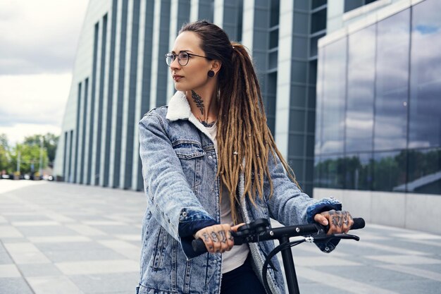 Atractiva chica elegante con rastas conduce una moto eléctrica en la calle cerca de un edificio interesante.