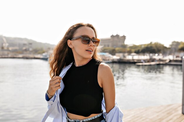 Atractiva chica encantadora con top negro y camisa azul con gafas de sol mirando a un lado con una sonrisa y disfrutando de caminar afuera en un día cálido y soleado