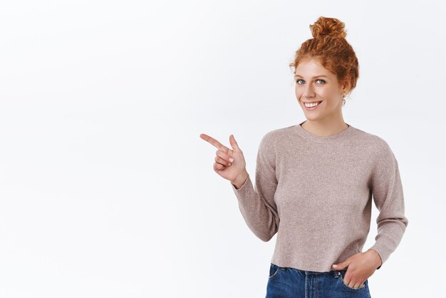 Atractiva chica con estilo con cabello rojo rizado en un moño desordenado usar suéter poner la mano en los bolsillos de los jeans apuntando a la izquierda anunciar promoción cámara sonriente mirarte como recomendar usar enlace fondo blanco