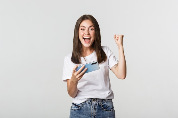 Atractiva chica morena alegre ganando en el juego para móviles, sosteniendo el teléfono inteligente y regocijándose.