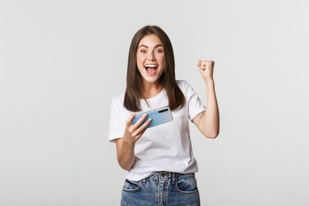 Atractiva chica morena alegre ganando en el juego para móviles, sosteniendo el teléfono inteligente y regocijándose.