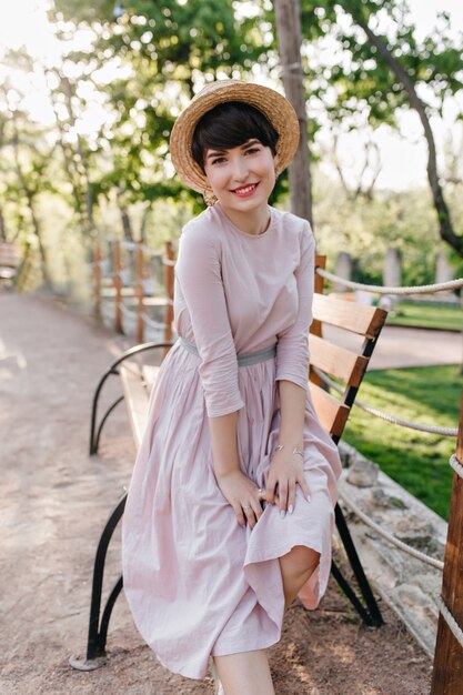 Atractiva chica morena inspirada en vestido largo y antiguo posando cerca del banco de madera y sonriendo