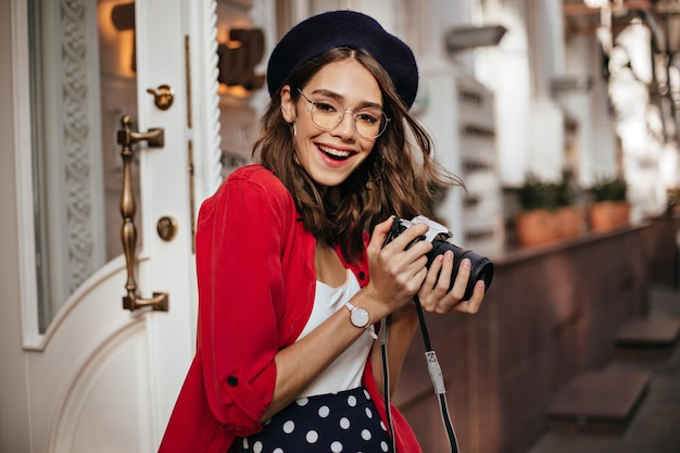 Atractiva chica pálida con cabello oscuro ondulado, labios rojos, boina de estilo francés, camisa roja y bonitos accesorios, sonriendo y haciendo fotos de la calle de otoño