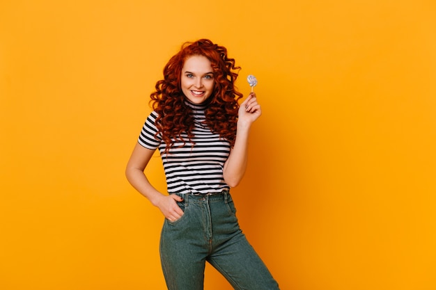 Atractiva chica pelirroja de ojos verdes en top recortada y jeans sonriendo y posando con piruleta en estudio naranja.