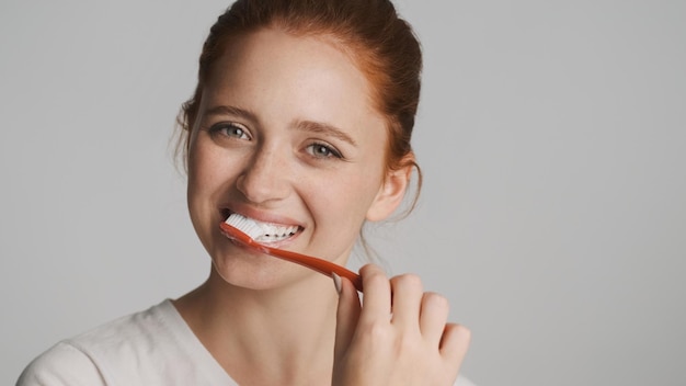 Atractiva chica pelirroja sonriente felizmente mirando en cámara cepillarse los dientes sobre fondo blanco.