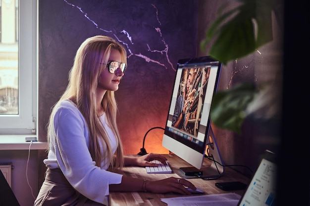 Atractiva chica rubia está estudiando en su habitación moderna usando una computadora.