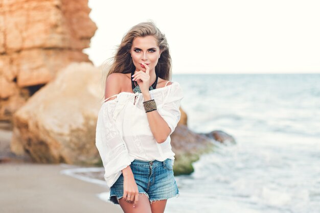 Atractiva chica rubia con pelo largo está posando en la playa cerca del mar. S Mantiene el dedo en los labios y sonríe a la cámara.