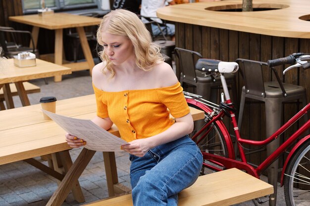 Atractiva chica rubia sentada en el patio del café mientras lee cuidadosamente el menú