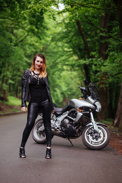 Foto gratuita una atractiva chica sexy vestida de cuero posando cerca de una motocicleta deportiva afuera