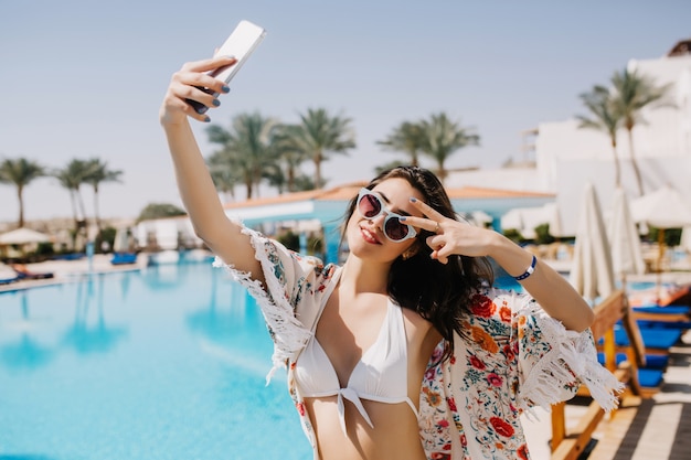 Atractiva chica sonriente divirtiéndose en el resort y haciendo selfie en paisaje sur con palmeras exóticas. Jovencita bronceada delgada en bikini blanco tomando una foto de sí misma mostrando el signo de la paz