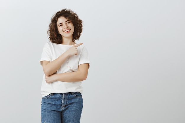 Atractiva chica sonriente mirando feliz y apuntando hacia la esquina superior derecha