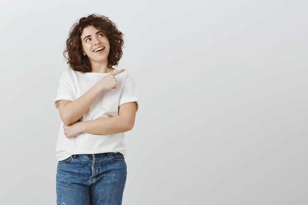 Atractiva chica sonriente mirando feliz y apuntando hacia la esquina superior derecha