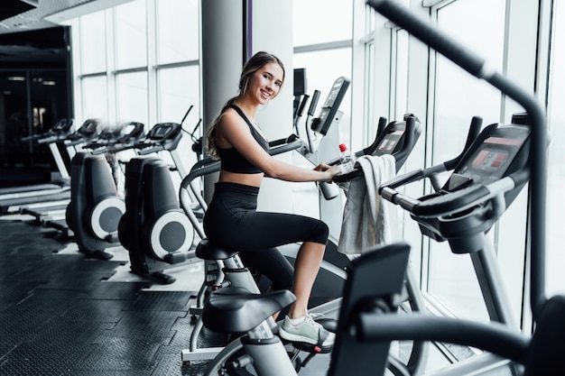 Atractiva chica sonriente onn bicicleta estática en un moderno y espacioso gimnasio