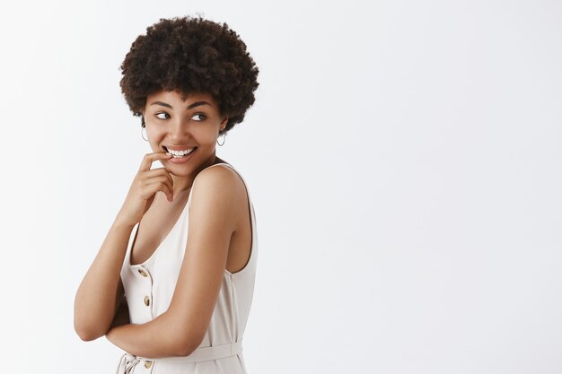 Atractiva y coqueta mujer afroamericana con peinado afro girando a la derecha con placer y deseo sonriendo, mordiéndose la uña con expresión curiosa e intrigada