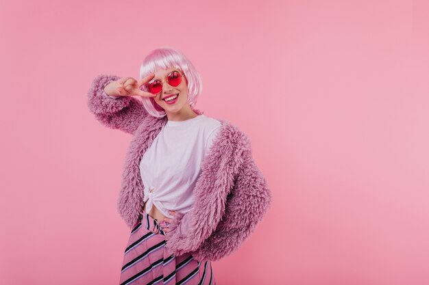 atractiva dama europea lleva elegantes gafas y peluca. Filmación en interiores de niña adorable sonriente en chaqueta de piel rosa