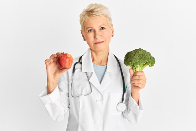 Atractiva doctora caucásica madura segura con manzana roja y brócoli verde como parte de una dieta saludable para reducir el riesgo de algunas enfermedades crónicas. Concepto de alimentación, nutrición y salud.