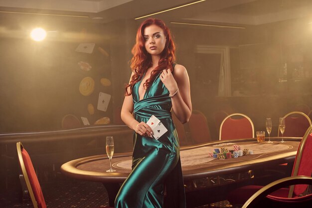 Atractiva doncella pelirroja con un vestido largo de satén azul posa de lado con dos ases en la mano contra una mesa de póquer en un casino de lujo. Pasión, cartas, fichas, alcohol, ganar, apostar: es como fem