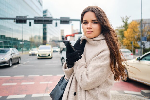 Atractiva y elegante chica morena con abrigo y teléfono celular mirando atentamente a la cámara caminando por las calles de la ciudad