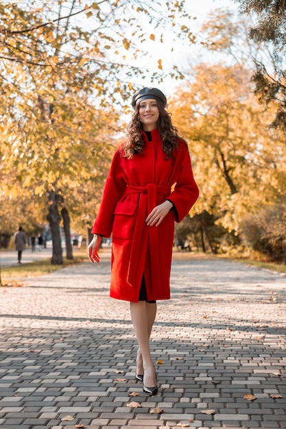 Atractiva y elegante mujer flaca sonriente con pelo rizado caminando en el parque vestida con abrigo rojo cálido otoño moda moda, estilo callejero, con sombrero boina