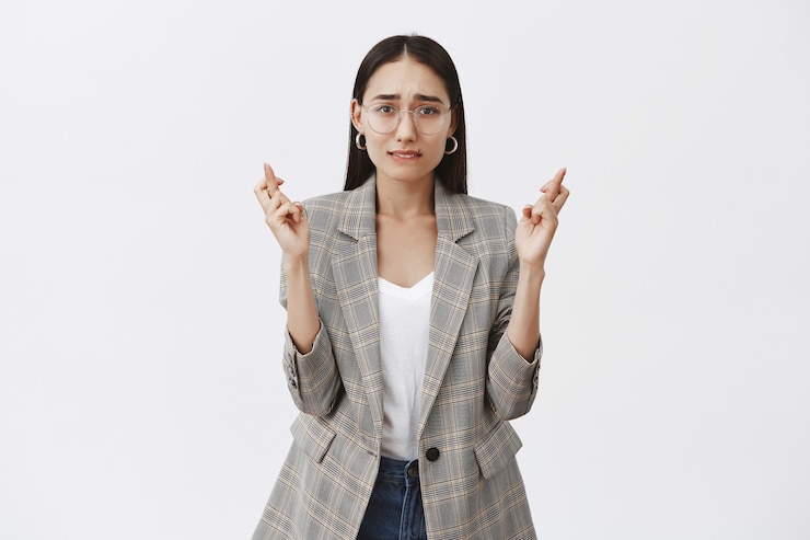 Foto gratuito atractiva empresaria adulta con gafas y chaqueta, cruzando los dedos y frunciendo el ceño mientras espera que el sueño se haga realidad.