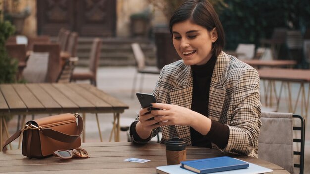 Atractiva empresaria alegre que parece feliz enviando mensajes de texto en un teléfono inteligente durante la pausa para el café en el patio de la cafetería