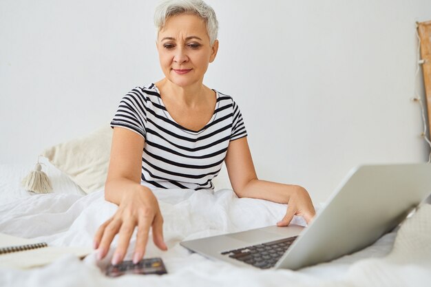 Atractiva empresaria anciana de pelo gris que trabaja de forma remota desde el dormitorio, sentada en la cama con una computadora portátil, usando la calculadora, administrando las finanzas, teniendo una expresión feliz y segura