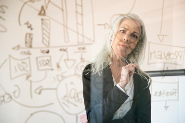 Atractiva empresaria mirando datos estadísticos y pensando. Gerente femenina pensativa experimentada confiada que sostiene el marcador y que se coloca en la sala de la oficina. Concepto de estrategia, negocios y gestión