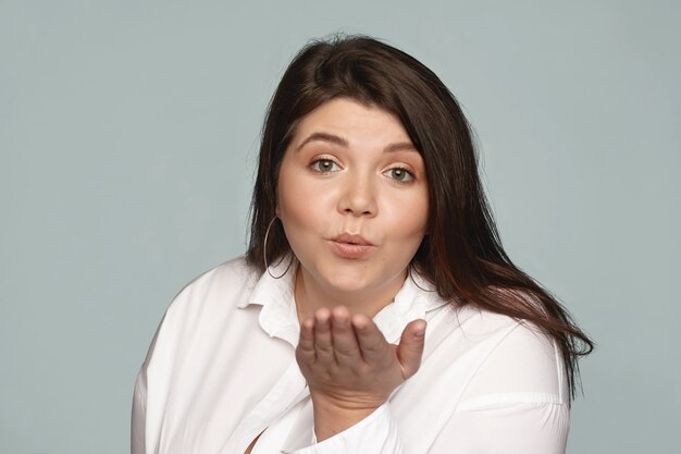 Atractiva y encantadora mujer joven coqueta con peso extra haciendo pucheros y enviándote amor y besos. Mujer juguetona gordita en camisa blanca soplando beso de aire