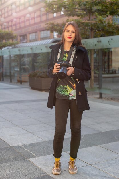 Atractiva fotógrafa turista con cámara, al aire libre en la calle de la ciudad. Hermosa mujer caucásica feliz en ropa casual hipster