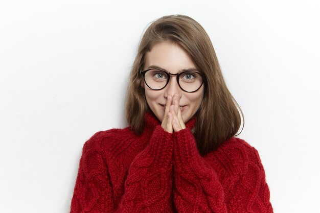 Atractiva joven alegre en gafas y suéter cálido y acogedor sosteniendo las manos juntas en su rostro y sonriendo felizmente, emocionado con noticias agradables, regalo, mirando