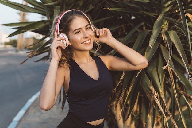 Atractiva joven alegre en ropa deportiva sonriendo, escuchando música a través de auriculares en la calle en una ciudad tropical. Modelo de moda, entrenamiento, entrenamiento, estado de ánimo alegre.