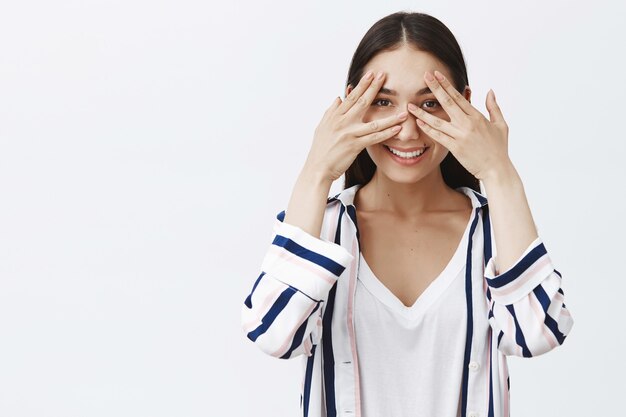 Atractiva joven encantadora y amigable con blusa a rayas, cubriéndose los ojos con las palmas y mirando a través de los dedos, sonriendo ampliamente