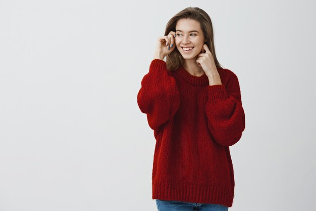 Atractiva joven estudiante adorando hermosos días nevados mirando por la ventana en suéter rojo suelto girando a la izquierda sonriendo satisfecho y tierno, tocando la piel suave y clara contra